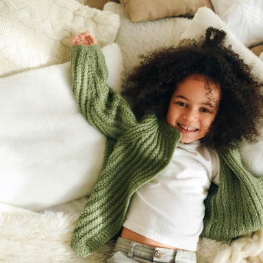 A young child dressed in a white shirt with a green sweater laying on a bed smiling up at the camera.