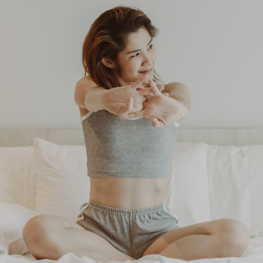A woman sitting on her made bed, stretching her arms out in front of her.