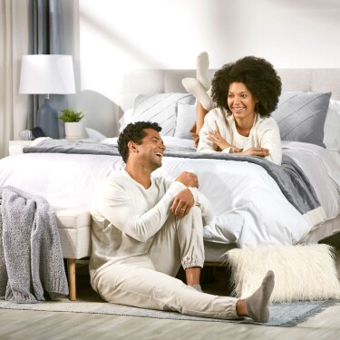 A man and a woman hanging out in a bedroom. The woman is laying across the bed while the man is sitting on a rug on the floor.
