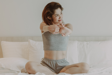 A woman sitting on her made bed, stretching her arms out in front of her.